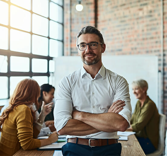 Cropped shot of a businessman standing in the office with his arms folded looking confident and smiling at the camera, in the background a team