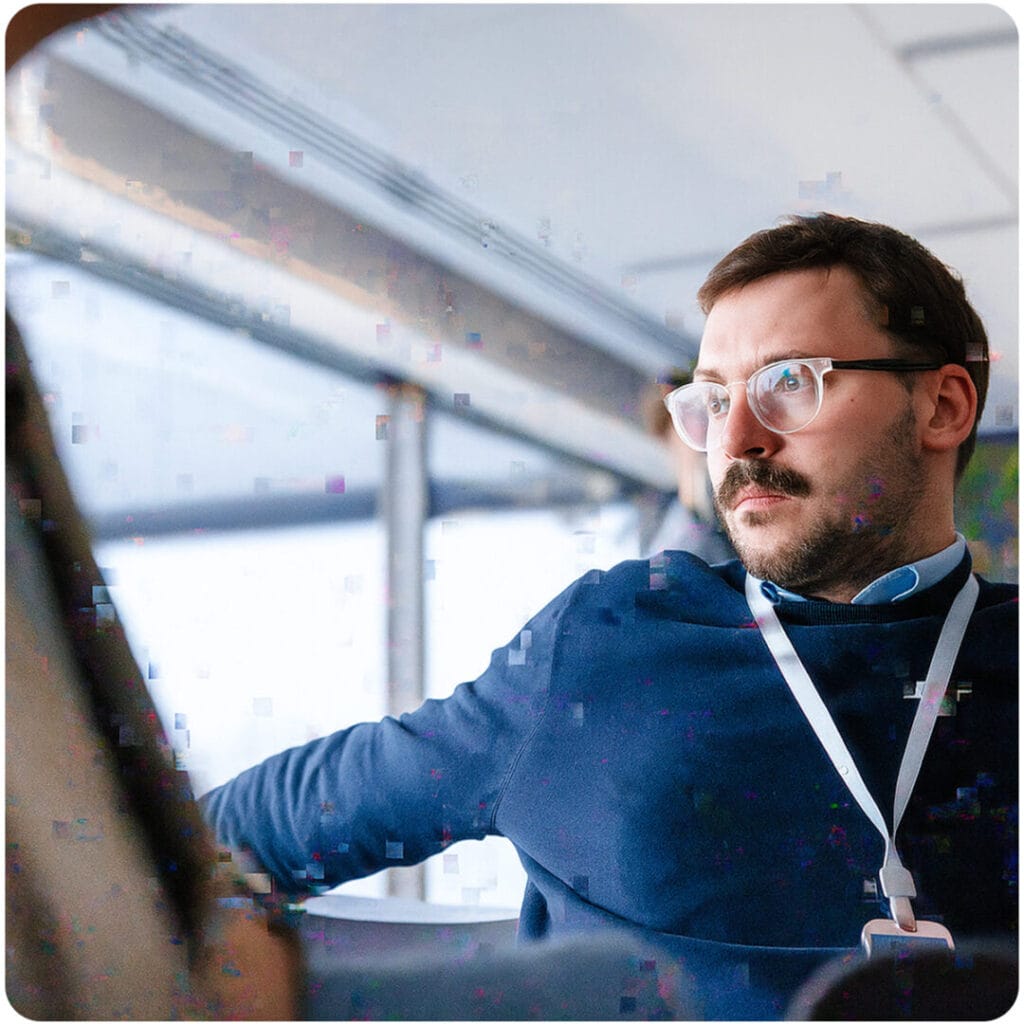 Mann mit Brille und Lanyard blickt nachdenklich aus dem Fenster, digitale Glitches auf dem Bild