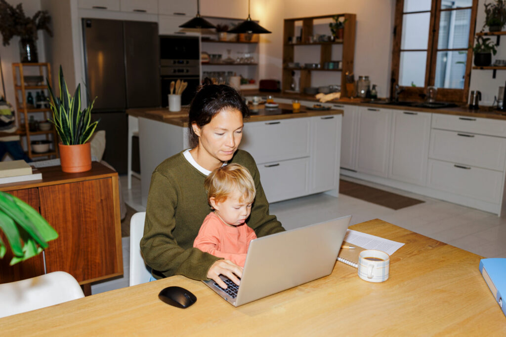 Mother multitasking, working remotely on laptop with baby while sitting at kitchen table
