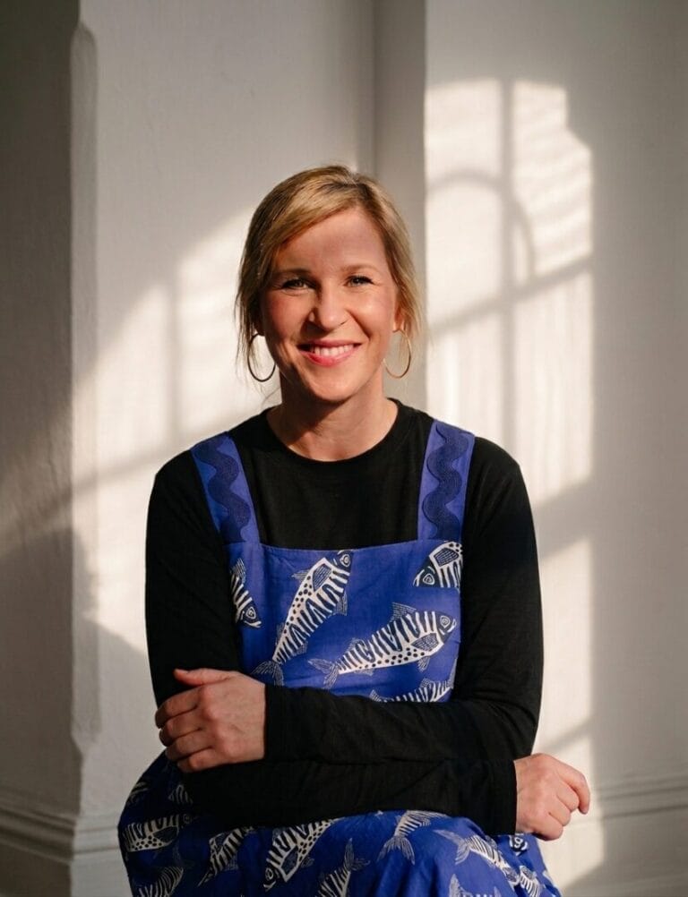 Portrait von Sophie Tretow, sitzend in einem blauen Kleid mit schwarz weißes Fischen drauf, Licht und Schatten fallen durch das Fenster