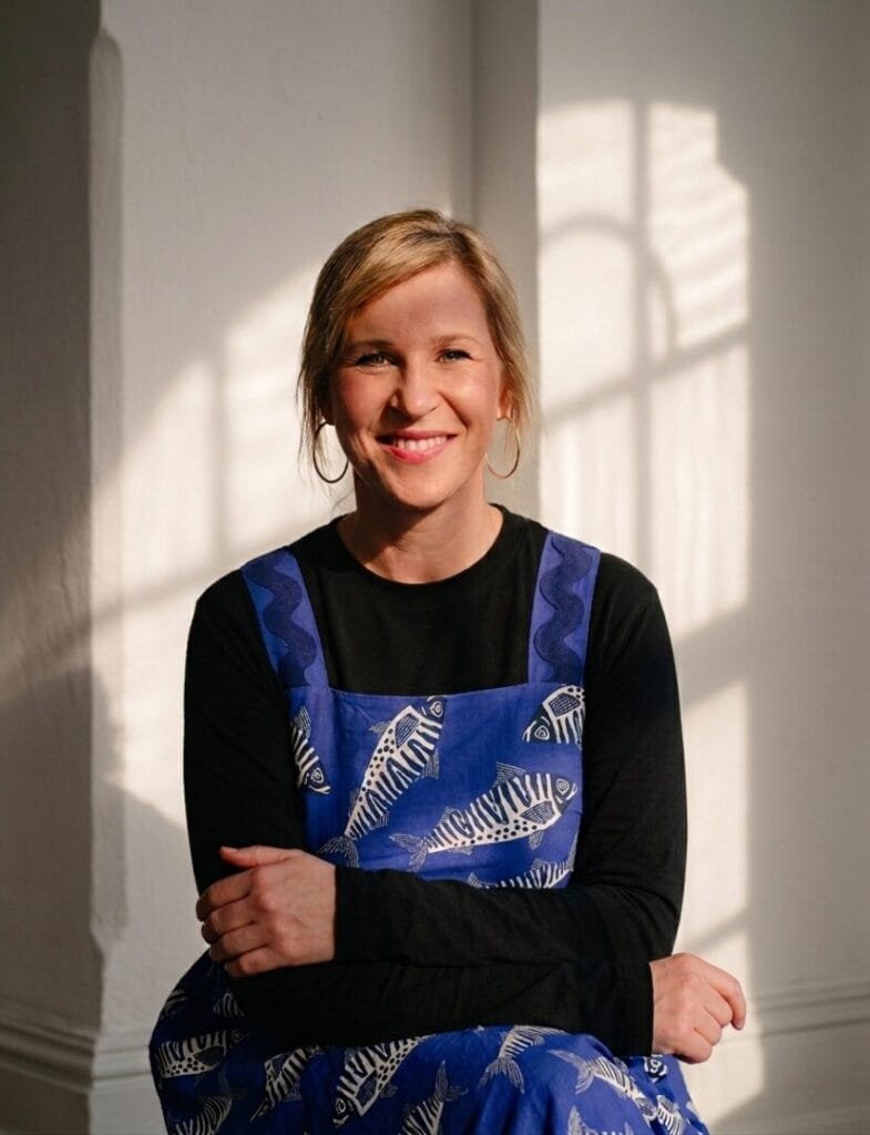 Portrait von Sophie Tretow, sitzend in einem blauen Kleid mit schwarz weißes Fischen drauf, Licht und Schatten fallen durch das Fenster